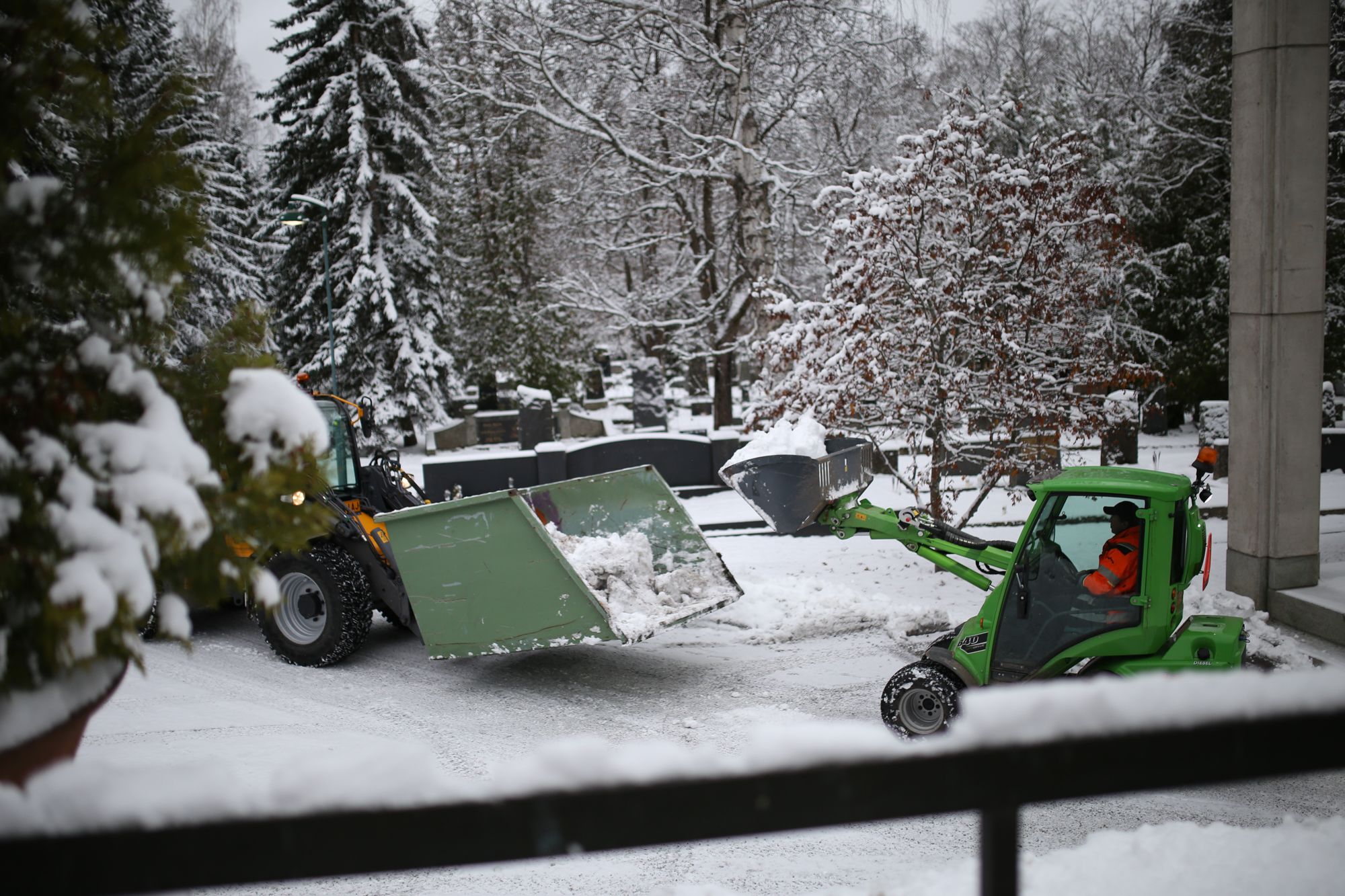 Pieni vihreä traktori kaataa lunta isomman traktorin kauhaan.