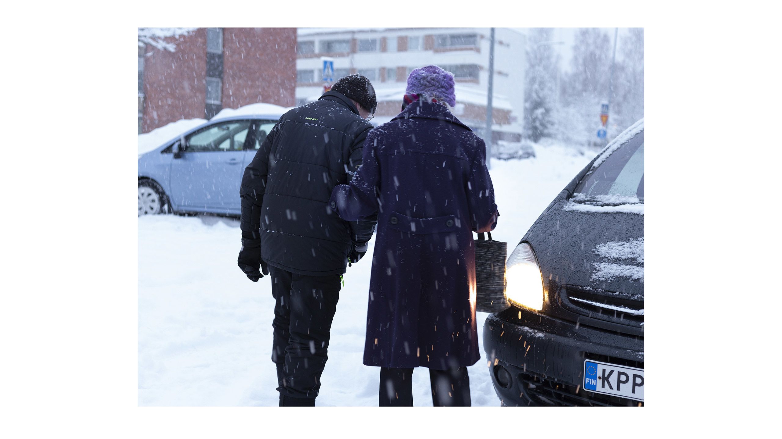 Ostoksilla olleet Saara Hentinen ja Jarmo Stolt poistuvat käsikynkkää ostoksilta. On paljon lunta ja Stolt saattaa iäkkään Hentisen autolle.