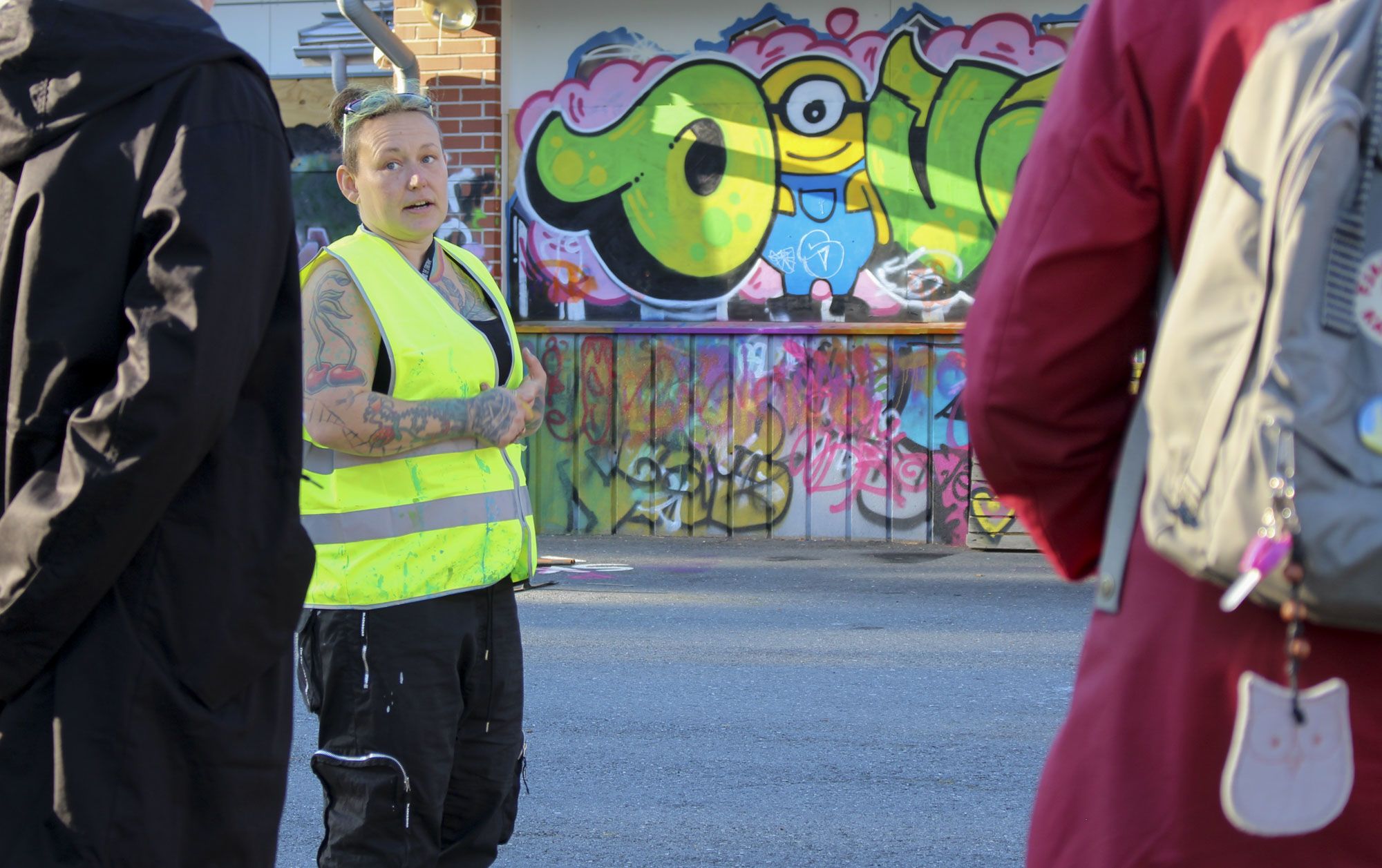 Piritta Suominen seisoo huomioliivissä ja puhuu kahdelle ihmiselle. Taustalla näkyy graffititaidetta.