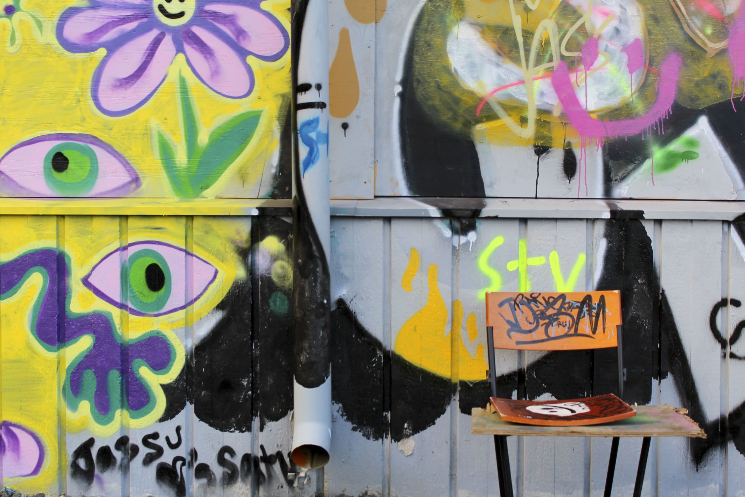 Seinällä on erilaisia graffitimaalauksia. Oikealla olevassa maalauksessa on violetteja silmiä. Seinän edessä on maaleilla töhritty tuoli.