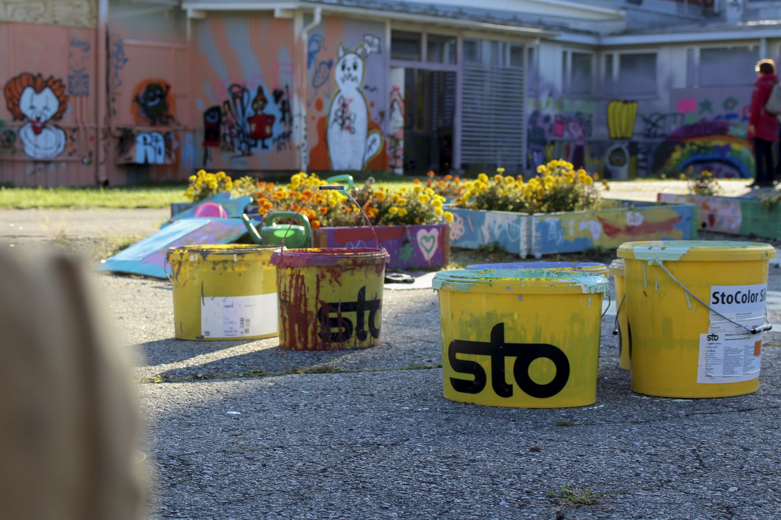 Maalipurkkeja asfaltilla. Taustalla on kukkia ja graffitimaalauksia.