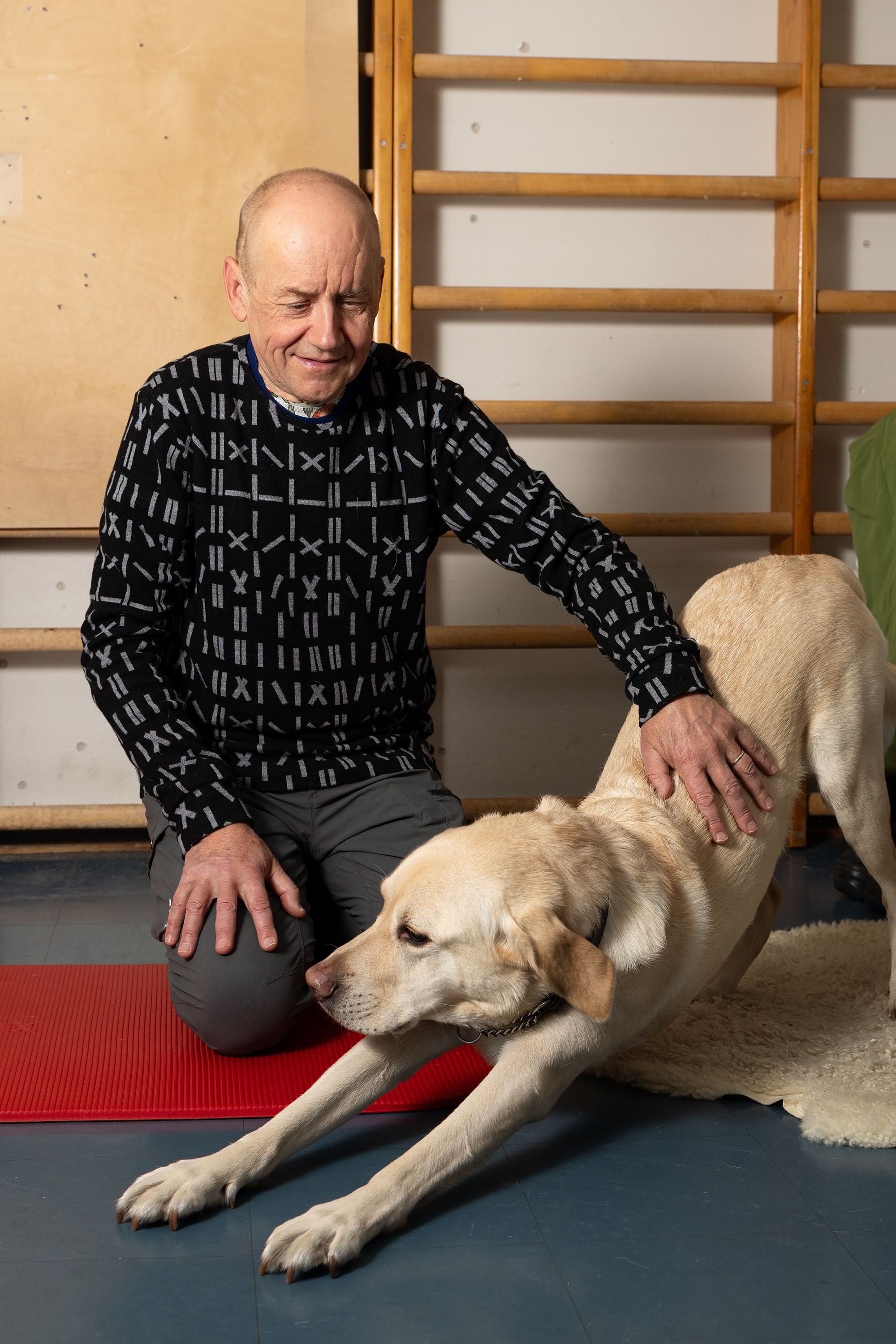 Janne Silas ja Patu-koira liikuntasalissa. Janne istuu lattialla ja silittää Patua hymyillen, Patu venyttelee.