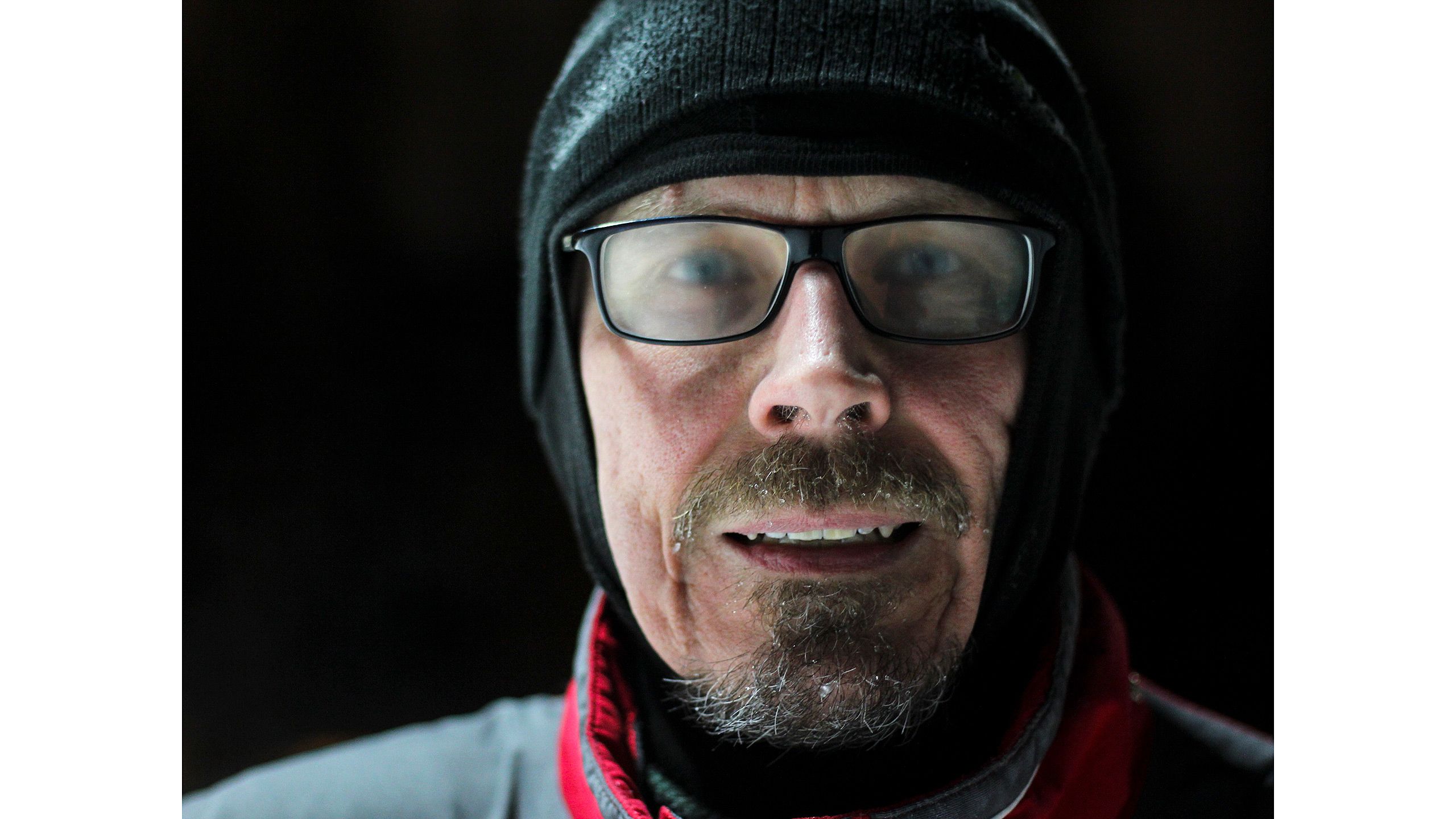 Lähikuva keski-ikäisen miehen kasvoista hiihtolenkin jälkeen. Miehen lasit ovat huurussa ja parta ja viikset kuurassa, ja hänellä on yllään musta pipo ja kypärämyssy.