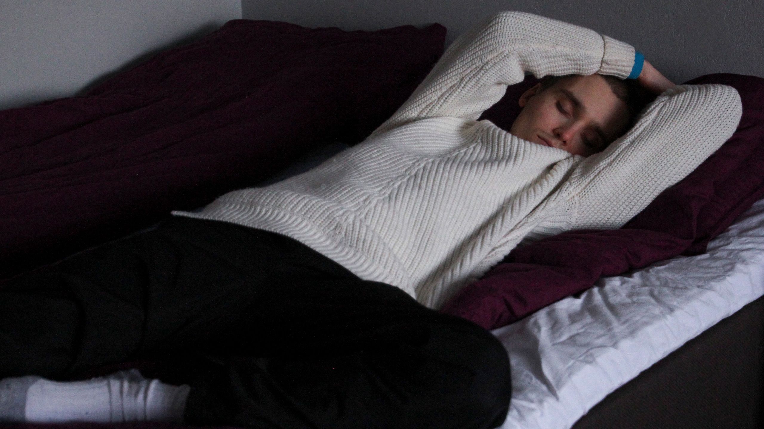 Valkopaitainen nuori mies nukkuu sängyllä peiton päällä.