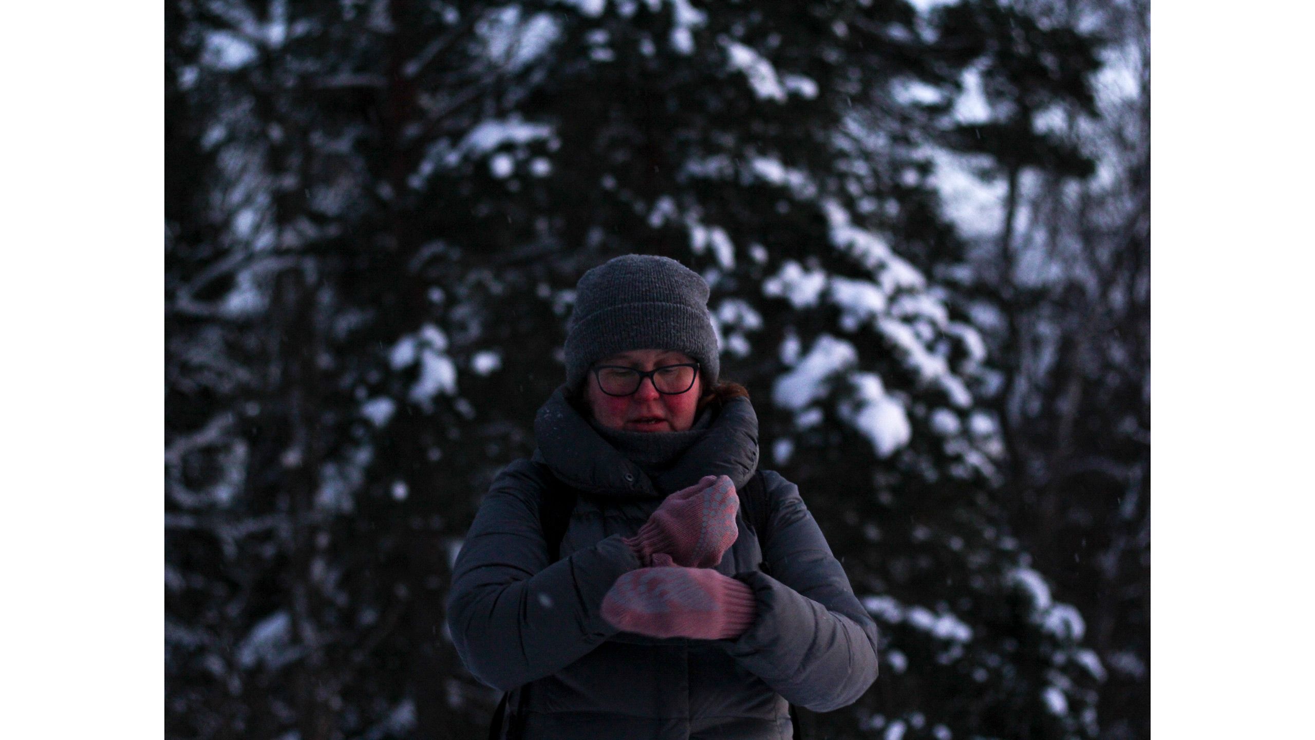 Nainen pukee roosanvärisiä lapasia ylleen. Hänen poskensa ja nenänpäänsä ovat pakkasen punoittamat. Taustalla näkyy luminen kuusi.