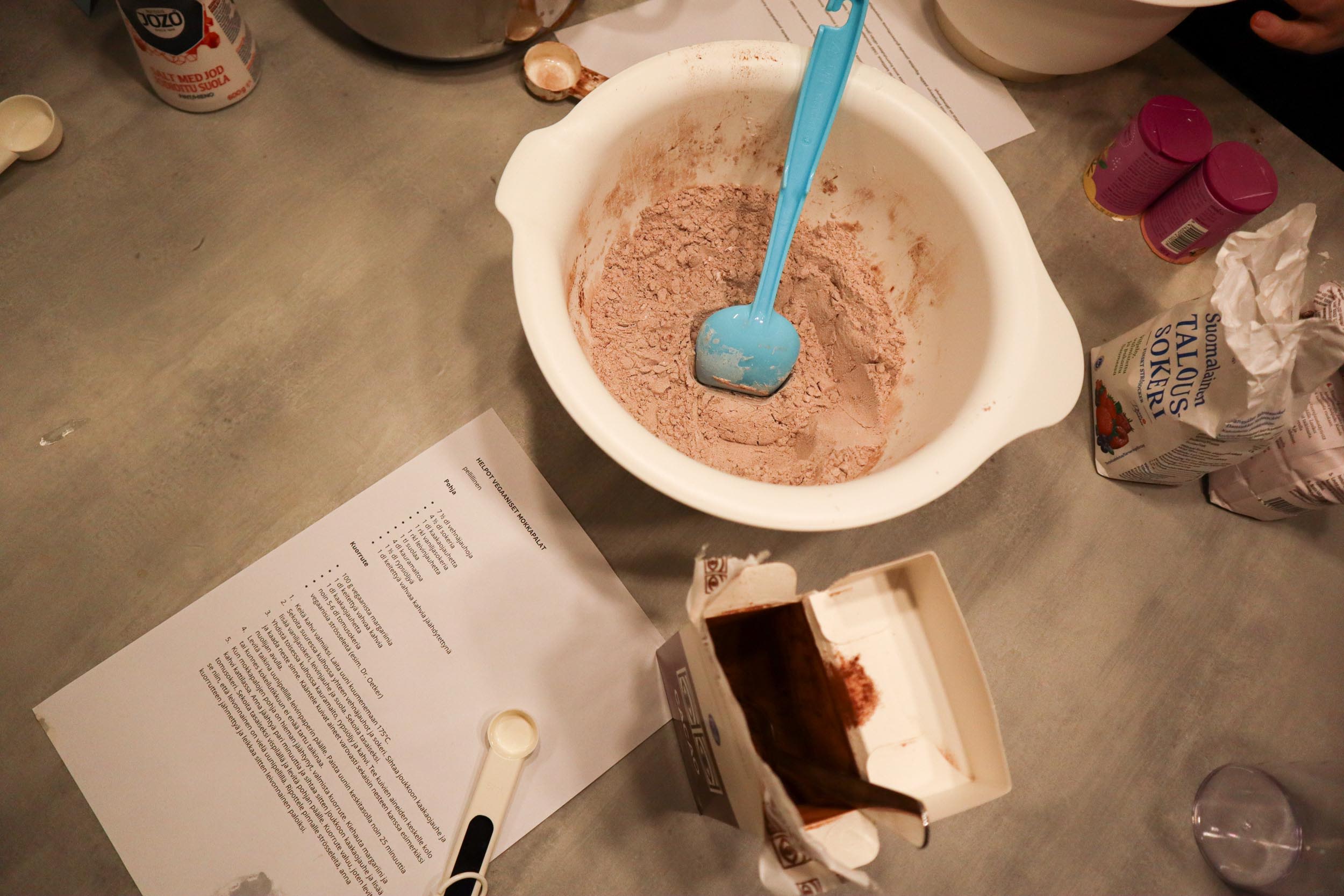 Kuvassa taikinakulho, leivontatarvikkeita ja mokkapalojen ohje.